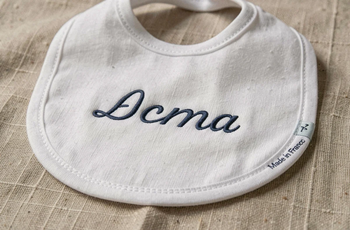 Bavoir bébé personnalisé en coton bio avec broderie prénom et label Made in France, posé sur du lin naturel